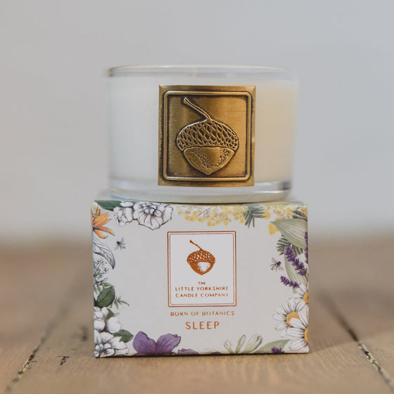 The Sleep travel candle is in a clear glass container with a copper acorn applique. The candle sits on top of its packaging, which features a delicate floral design.