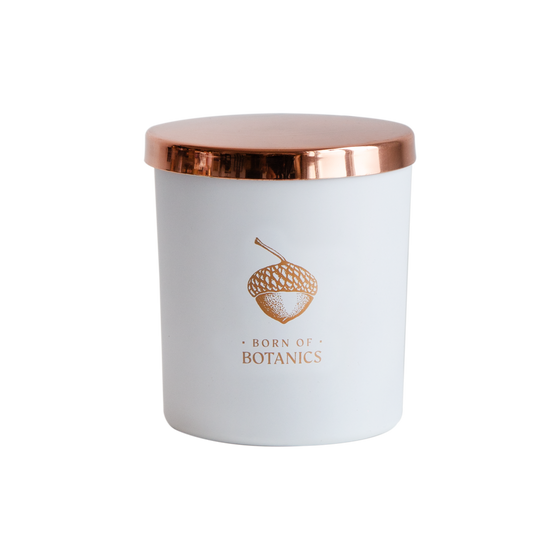 A cut-out image of the Serenity candle. It is in a matte-white container with a copper acorn logo, underneath which "Born of Botanics" is printed in copper.  It is topped with a shiny copper lid.